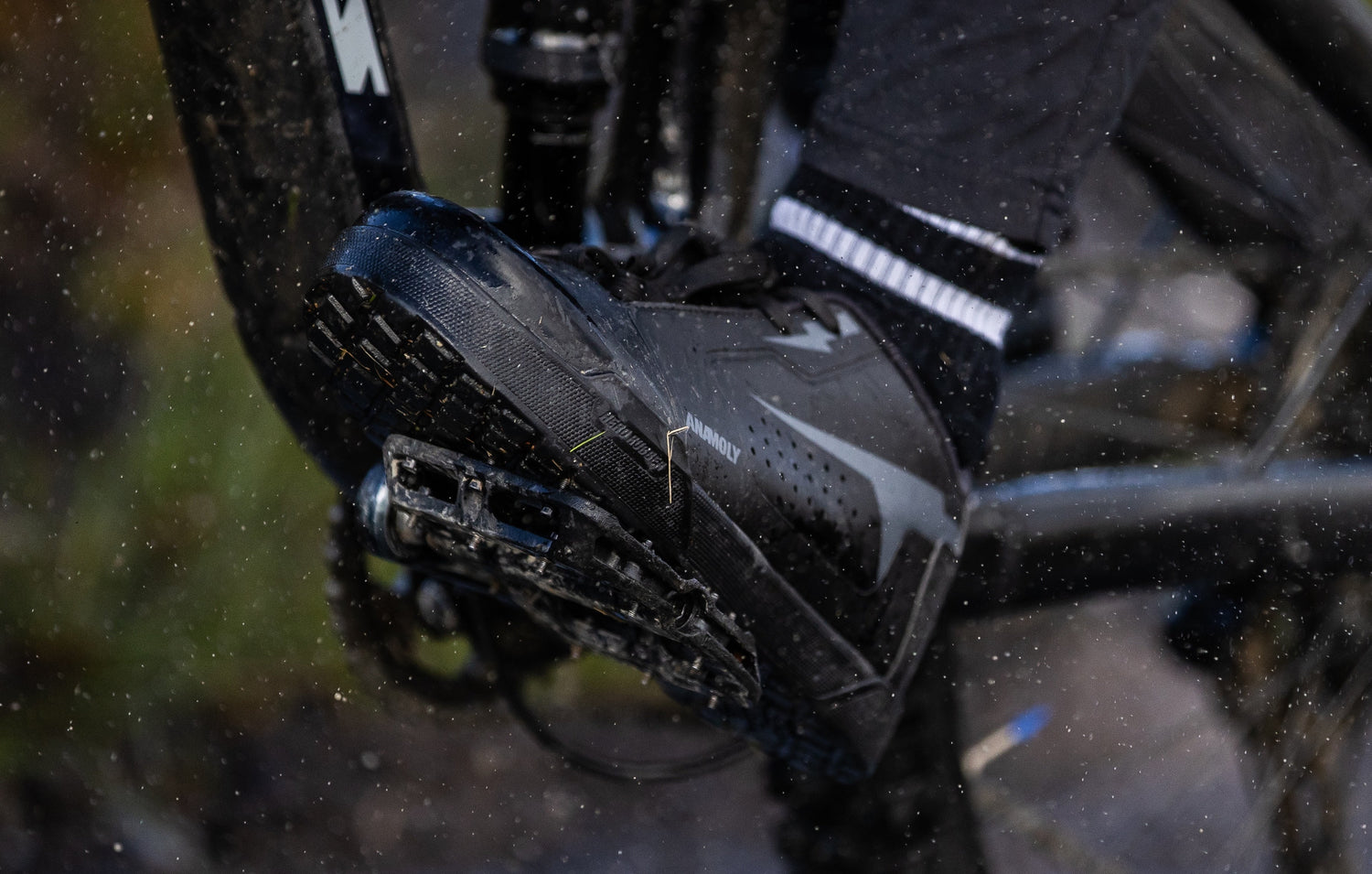 Macro close-up of MTB shoe sole with textured rubber compound gripping metal pedal pins on a wet rocky surface